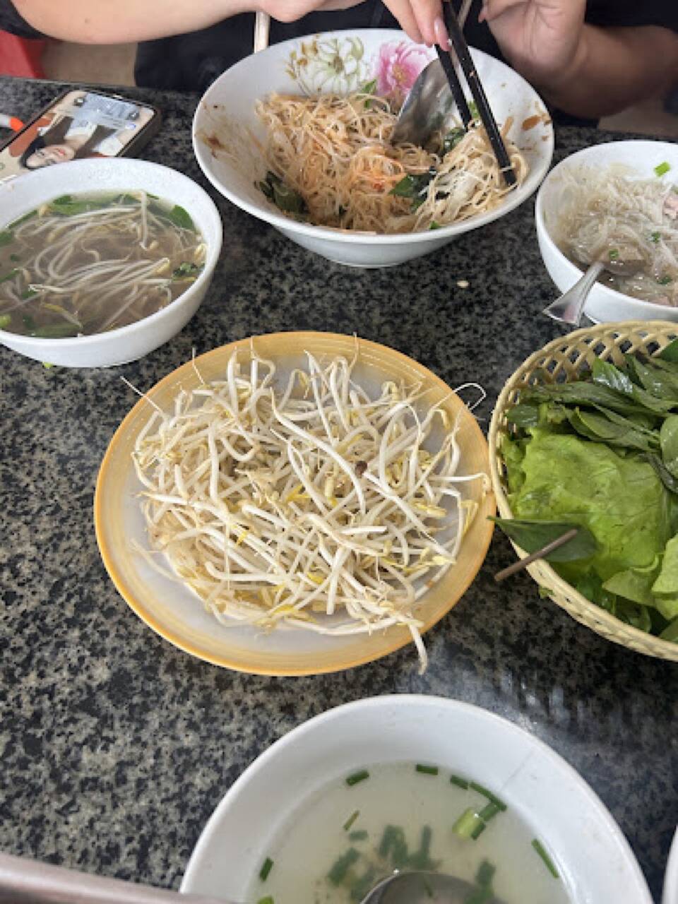Bàn ăn tại Phở Ngọc Sơn với đĩa giá đỗ, rổ rau và các bát nước dùng