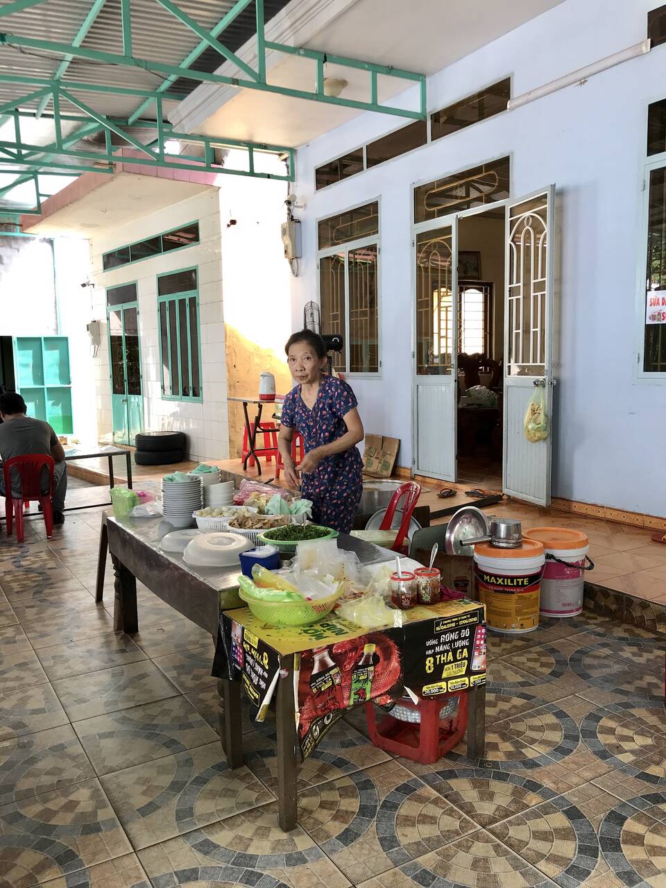 Quầy bánh canh ngoài hiên nhà của Bánh Canh Cô Bông, chủ quán đứng sau bàn bày chén và rau