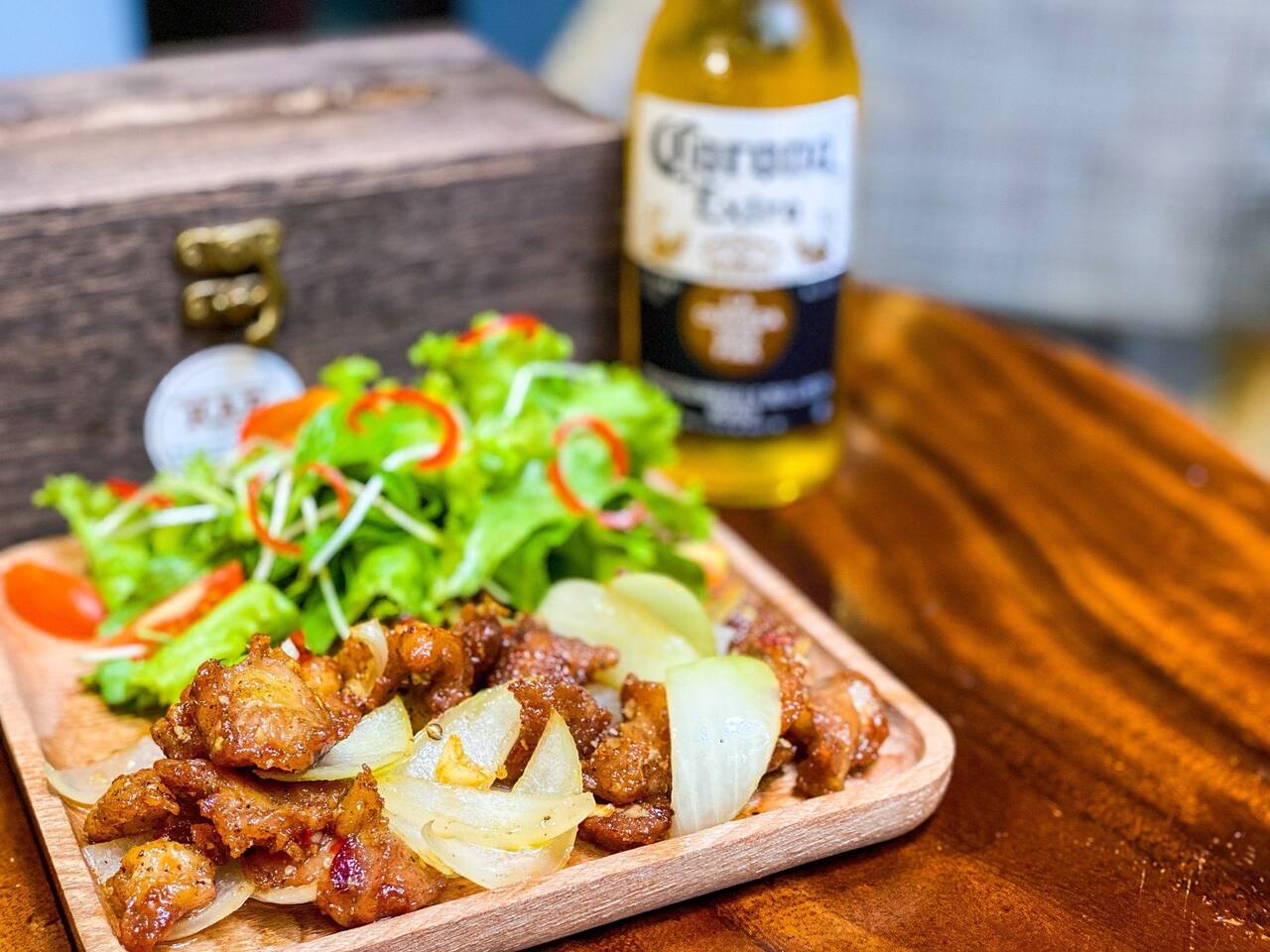 Đĩa thịt chiên giòn kèm rau và chai bia trên khay gỗ tại KAP Bistro