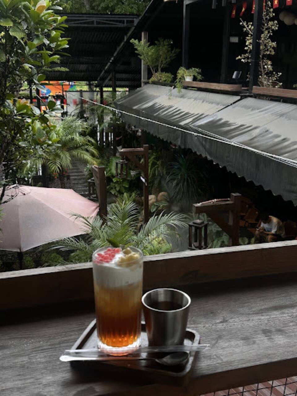 Ly trà trái cây phủ kem đặt trên bàn ngoài hiên quán The Quao, nhiều cây xanh phía sau