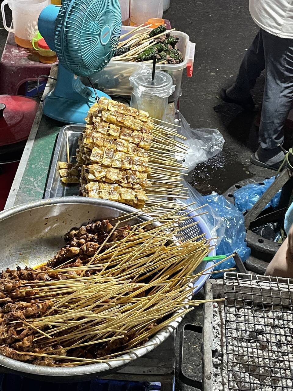 Xiên thịt xếp trên khay và chảo, cạnh bếp nướng ở một quầy trong chợ đêm