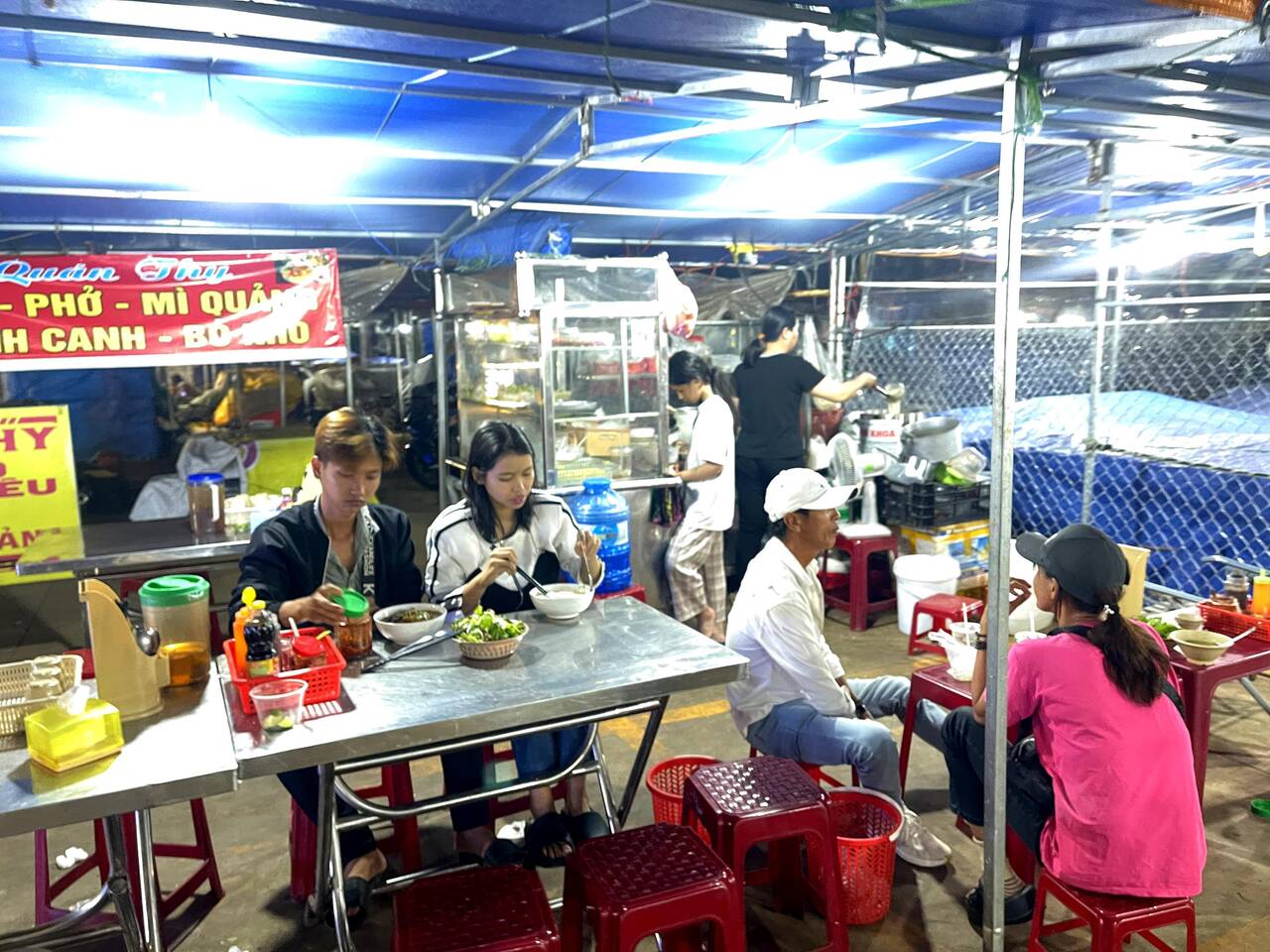 Hàng phở, mì, bánh canh dưới lều xanh với bàn inox và ghế đỏ, khách ngồi ăn