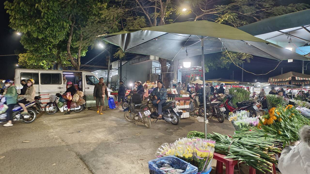 Hoa tươi và rau xếp trên nền đất ở chợ ban đêm, xe máy đỗ phía sau