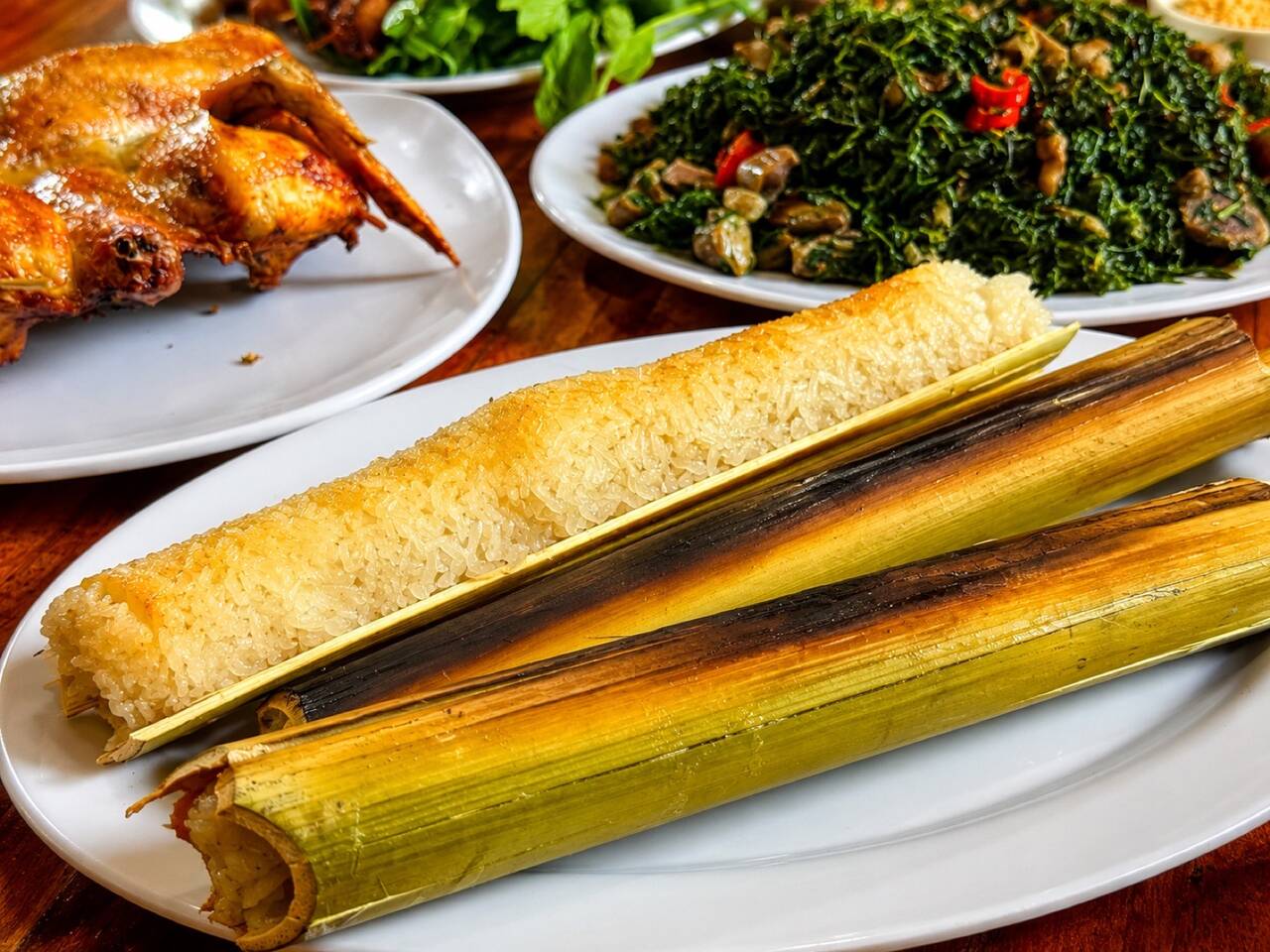 Đĩa gà nướng, ống cơm lam nướng vàng và đĩa rau rừng xào tại Bazan
