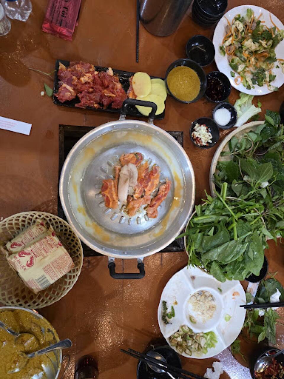 A pan of marinated meat grilling beside a basket of fresh greens and dipping sauce at Quán Nhà Tôi