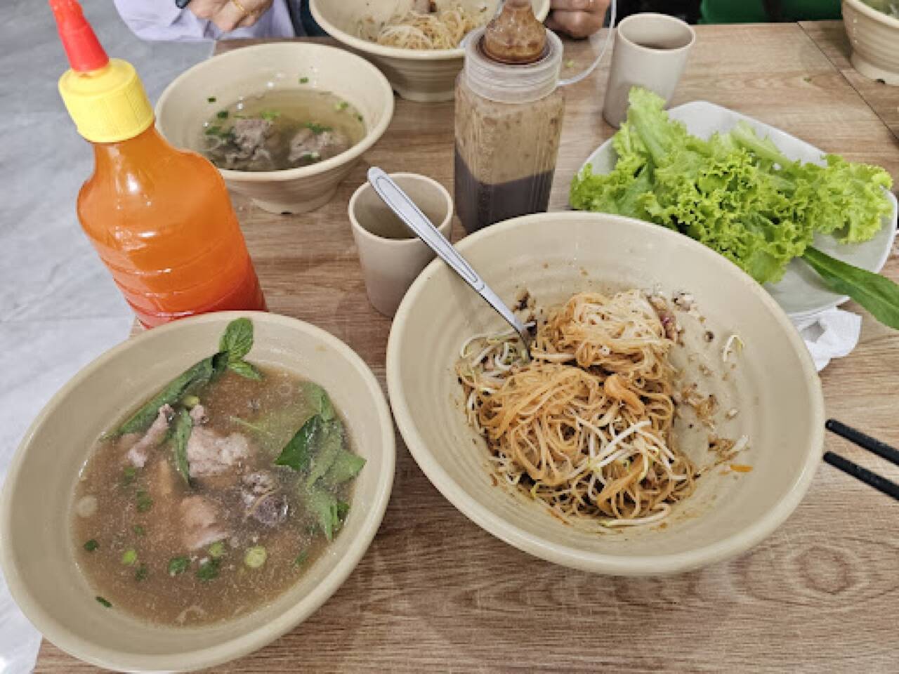 Phở khô trộn sốt kèm bát canh xương, đĩa xà lách và chai tương ớt trên bàn gỗ tại Phở khô Minh