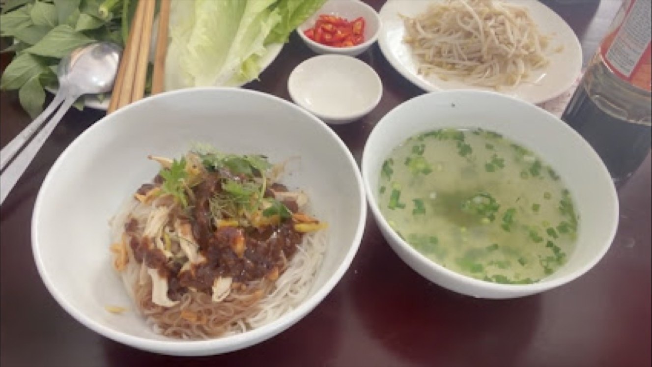 Bát phở khô trộn thịt và bát nước dùng có bánh phở, rau xà lách, ớt và tương đặt trên bàn tại Phở Nhớ Phố Núi