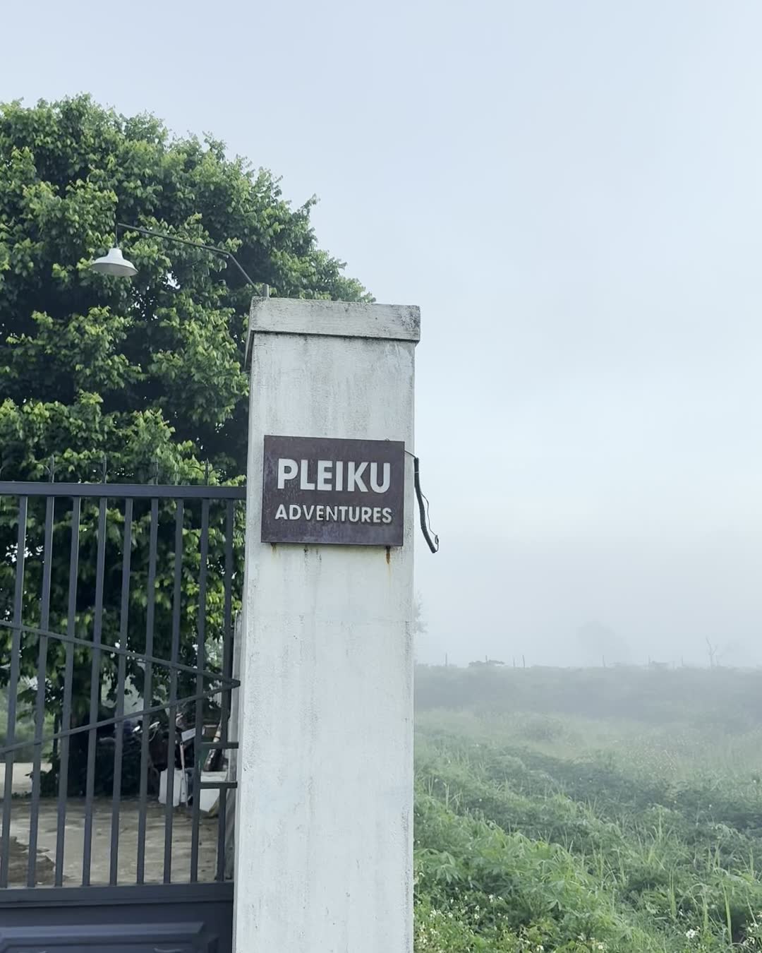 Cột cổng có biển Pleiku Adventures, phía sau là cánh đồng trong sương sớm