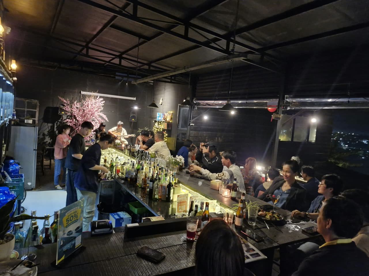 Quầy bar dài đông khách, phía sau có người chơi guitar hát nhạc sống
