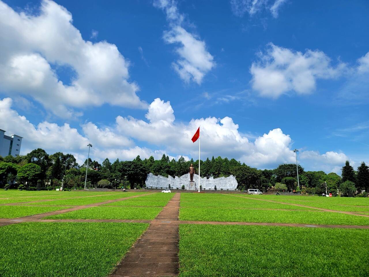 Bãi cỏ chia ô vuông bằng lối gạch, cột cờ đỏ trên cột trắng cao và tượng trước bức tường phù điêu xám