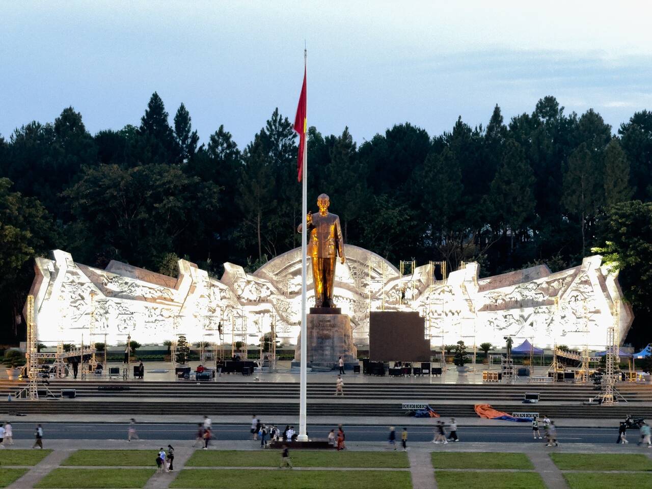 Tượng vàng trước bức tường phù điêu trắng trải dài hai bên, cột cờ đỏ cao phía trước